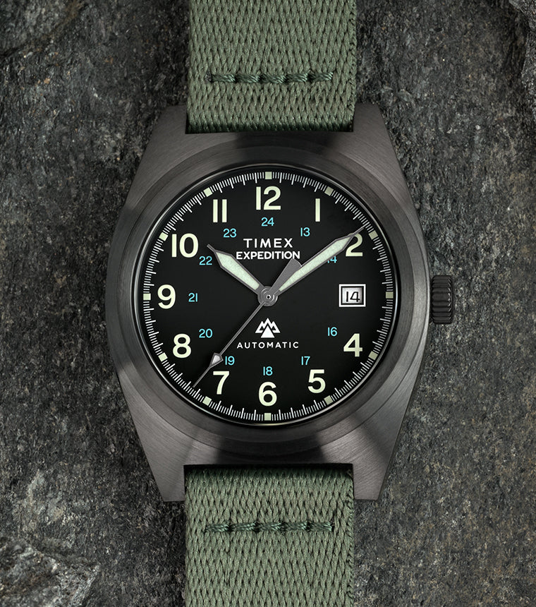 Timex Expedition Capstone Automatic field watch with black dial, luminous Arabic numerals, date window at 3 o’clock, and green fabric strap, displayed on rugged stone background.
