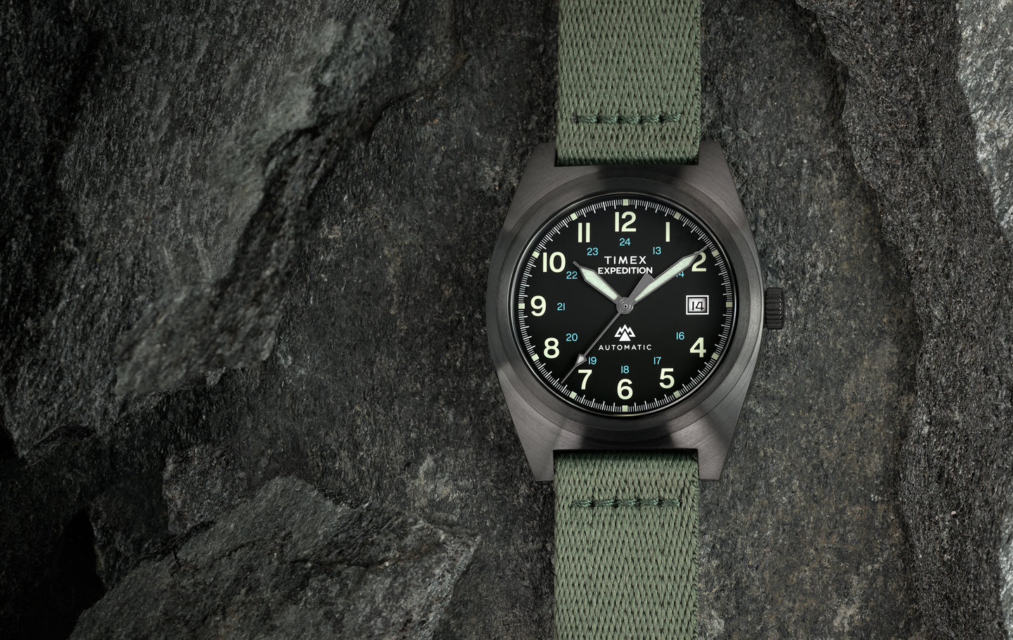 Timex Expedition Capstone Automatic field watch with black dial, luminous Arabic numerals, date window at 3 o’clock, and green fabric strap, displayed on rugged stone background.