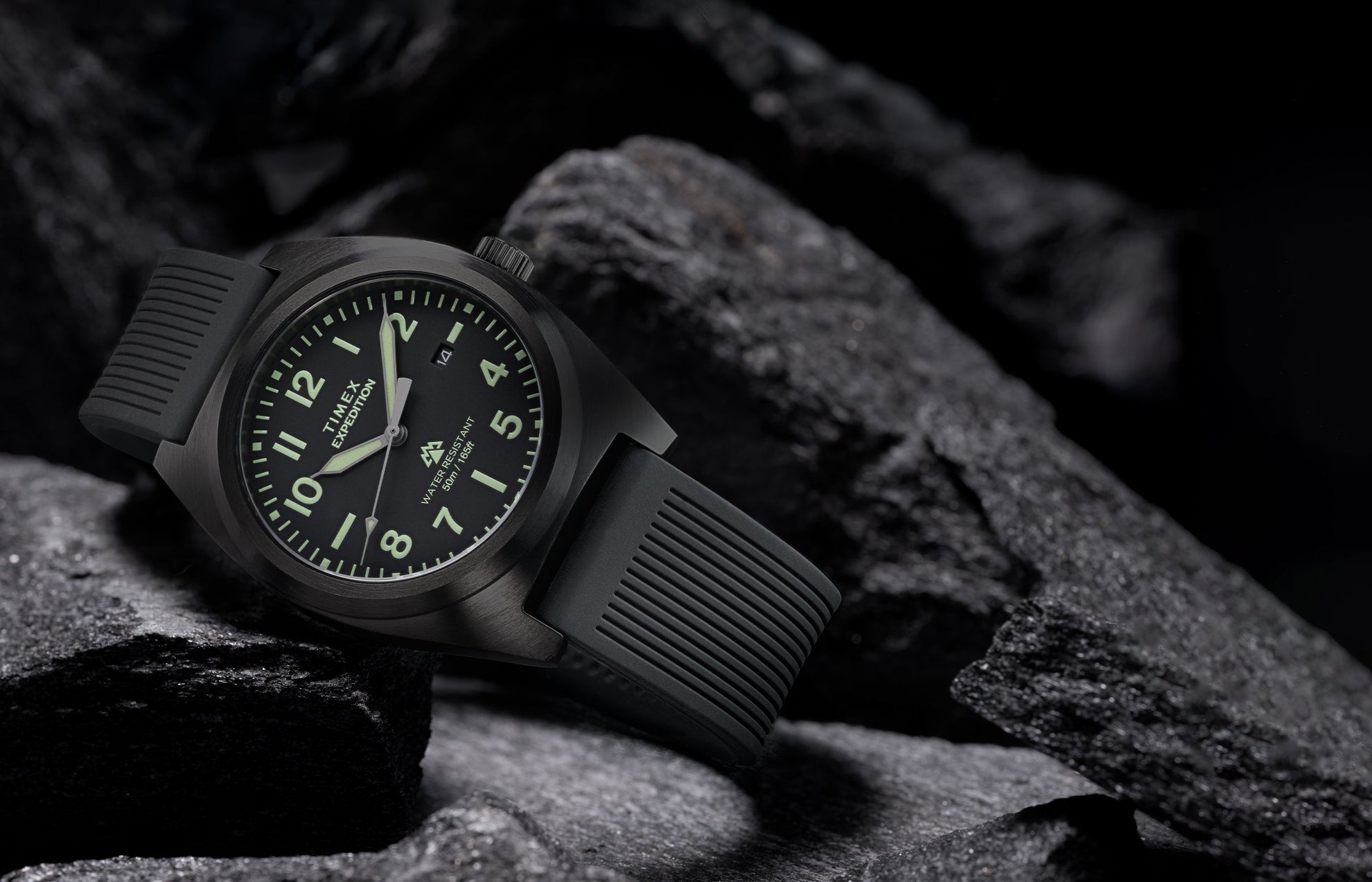 Timex Expedition® Capstone watch with black dial and black silicone strap resting among rugged dark rocks. The watch face glows softly with luminous markers, highlighting its durability, 50-meter water resistance, and adventure-ready design.