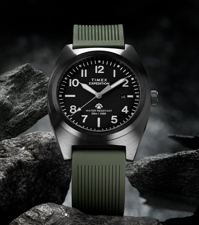 Timex Expedition® Capstone watch with a black dial and olive green silicone strap, displayed among rugged dark rocks. The watch face shows bold white numerals and hands, highlighting its adventure-ready design and 50-meter water resistance.