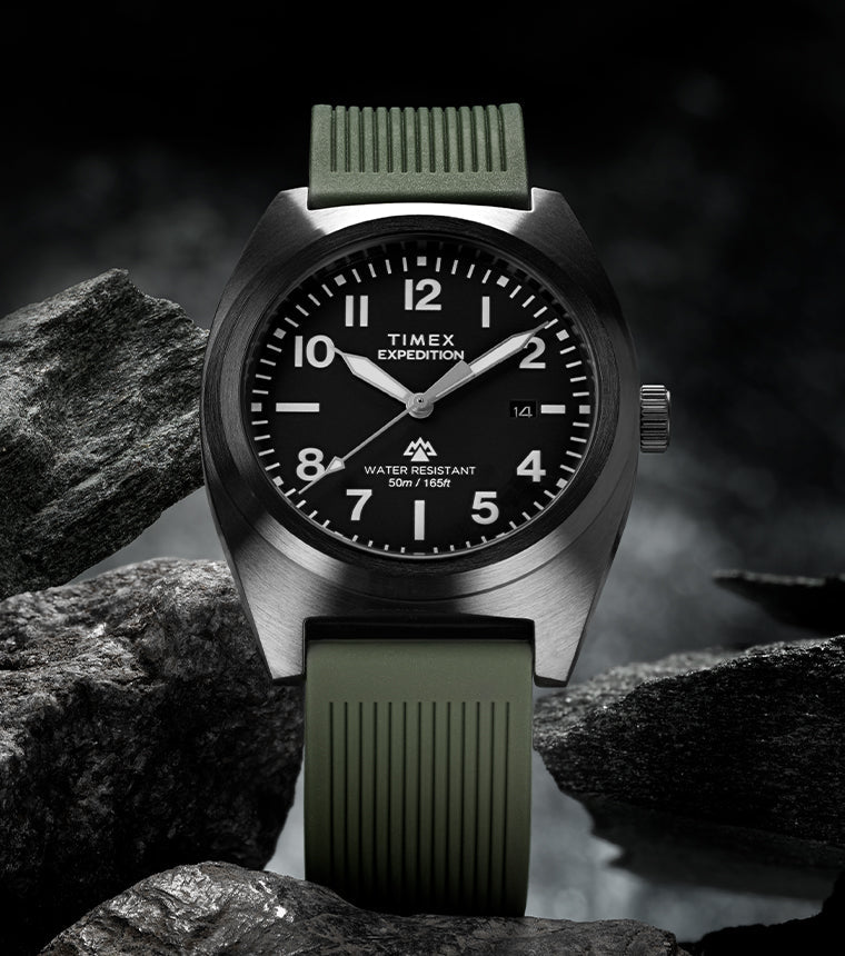 Timex Expedition® Capstone watch with a black dial and olive green silicone strap, displayed among rugged dark rocks. The watch face shows bold white numerals and hands, highlighting its adventure-ready design and 50-meter water resistance.