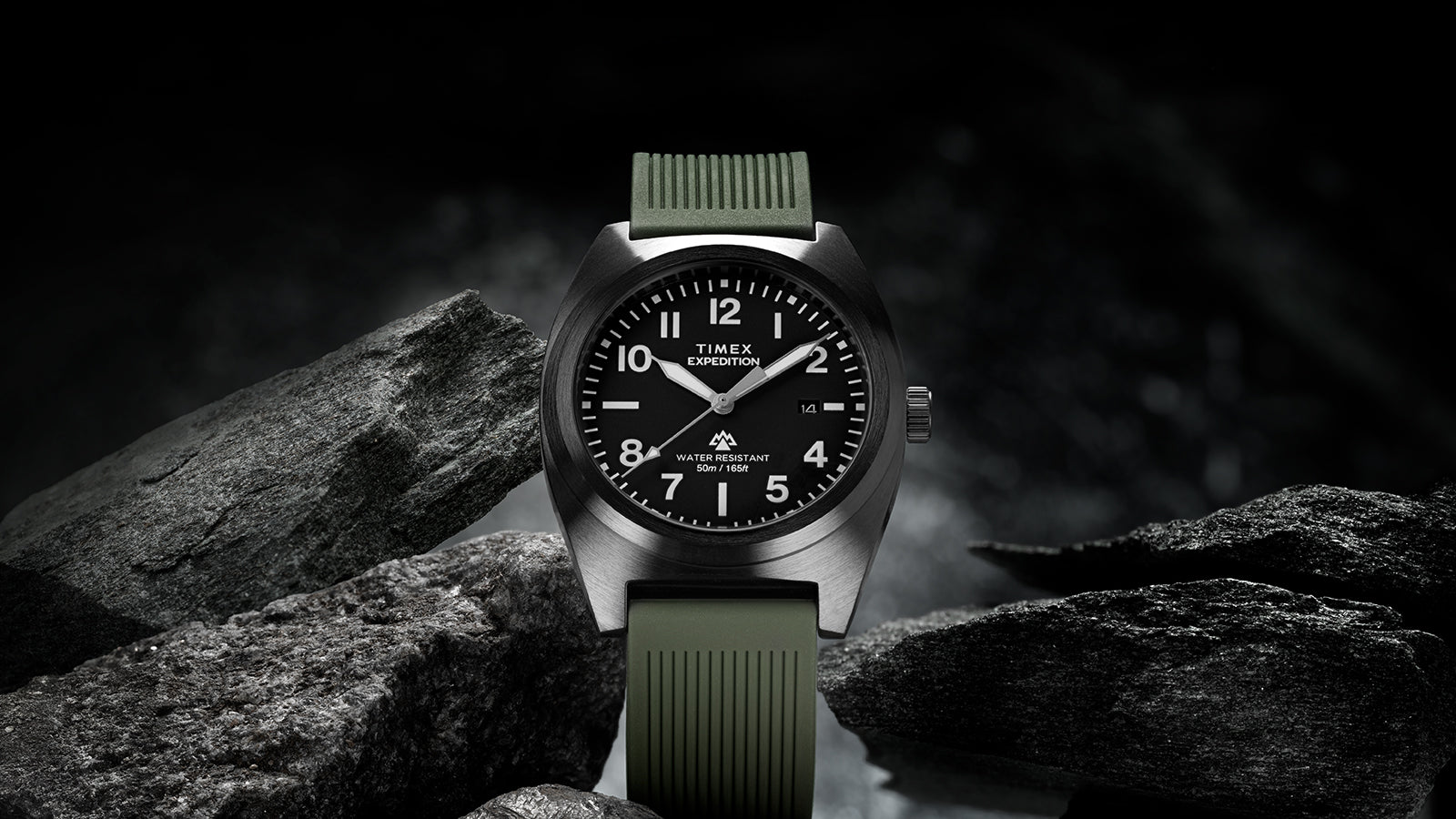 Timex Expedition® Capstone watch with a black dial and olive green silicone strap, displayed among rugged dark rocks. The watch face shows bold white numerals and hands, highlighting its adventure-ready design and 50-meter water resistance.