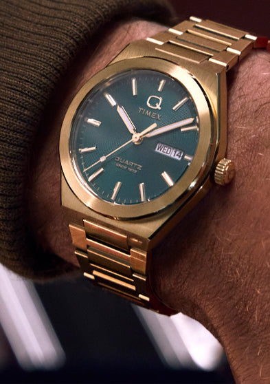 Wrist shot of the gold-tone Q Timex® Continental Day and Date watch with green dial, seen from an angle. The brushed bracelet and bold dial stand out in a relaxed setting, reflecting a retro-inspired luxury look.