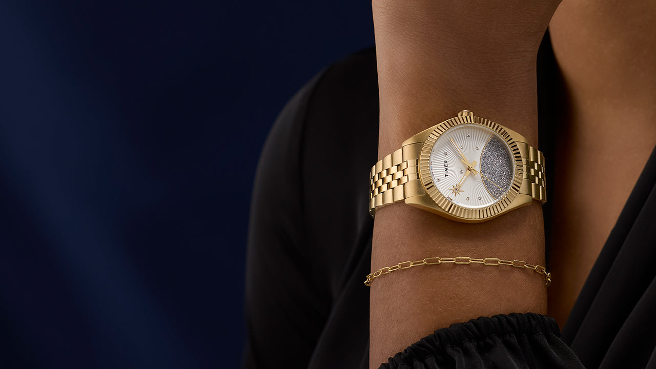 Timex Legacy gold-tone watch styled on wrist with a black sleeve and gold chain bracelet, showcasing its elegant crystal-accented dial and textured glitter detail.