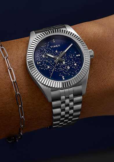 Timex Legacy watch worn on a wrist, featuring a blue starry dial and silver bracelet paired with a minimalist chain bracelet for an elegant look.