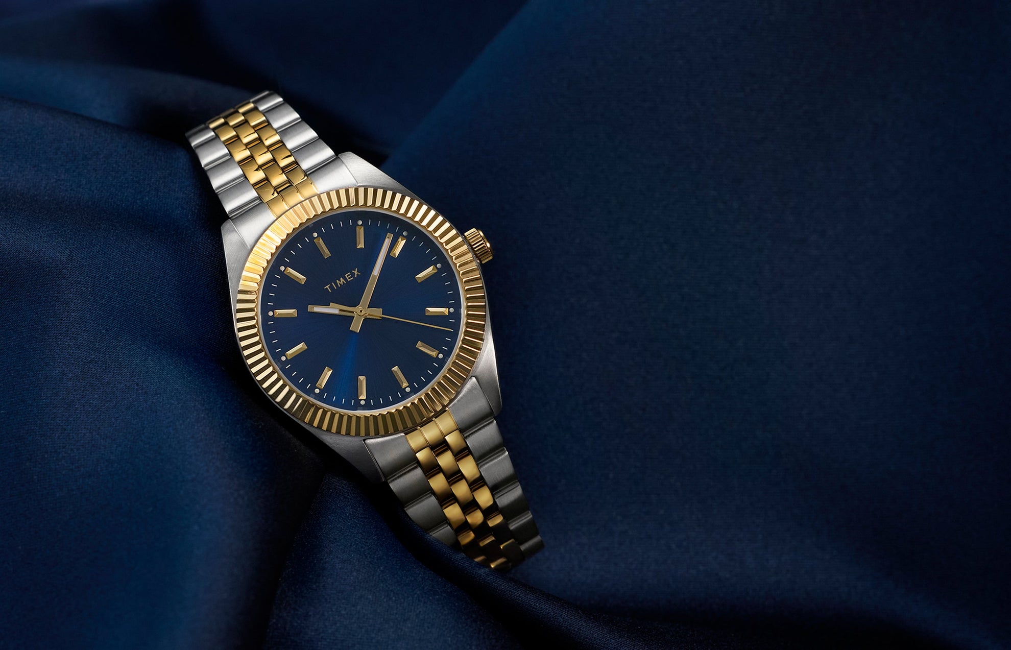 Timex Legacy watch with a blue dial, gold-tone markers, and a two-tone stainless-steel bracelet, displayed on deep blue satin fabric for an elegant look.