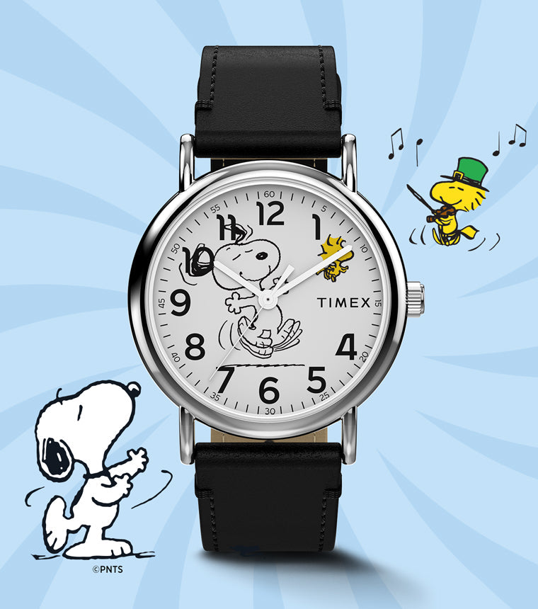 Timex x Peanuts® Weekender watch with a white dial featuring Snoopy dancing with Woodstock, displayed on a light blue background with playful Peanuts® illustrations. The watch has a black leather strap and classic silver-tone case.