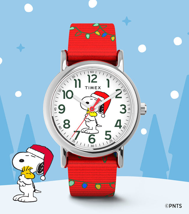 Timex x Peanuts® Weekender Snoopy Santa Hat watch featuring Snoopy in a Santa hat holding Woodstock on a white dial with green numerals. The watch has a red fabric strap decorated with holiday string lights, set against a snowy winter background with Snoopy and Woodstock illustration.