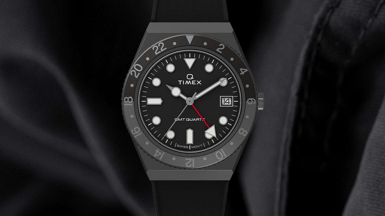 Front view of the Q Timex GMT watch in matte black finish, featuring a black dial with white circular and triangular hour markers, a red GMT hand, and a date window at 3 o’clock. The bezel shows 24-hour markings in grey, and the watch is paired with a black rubber strap.