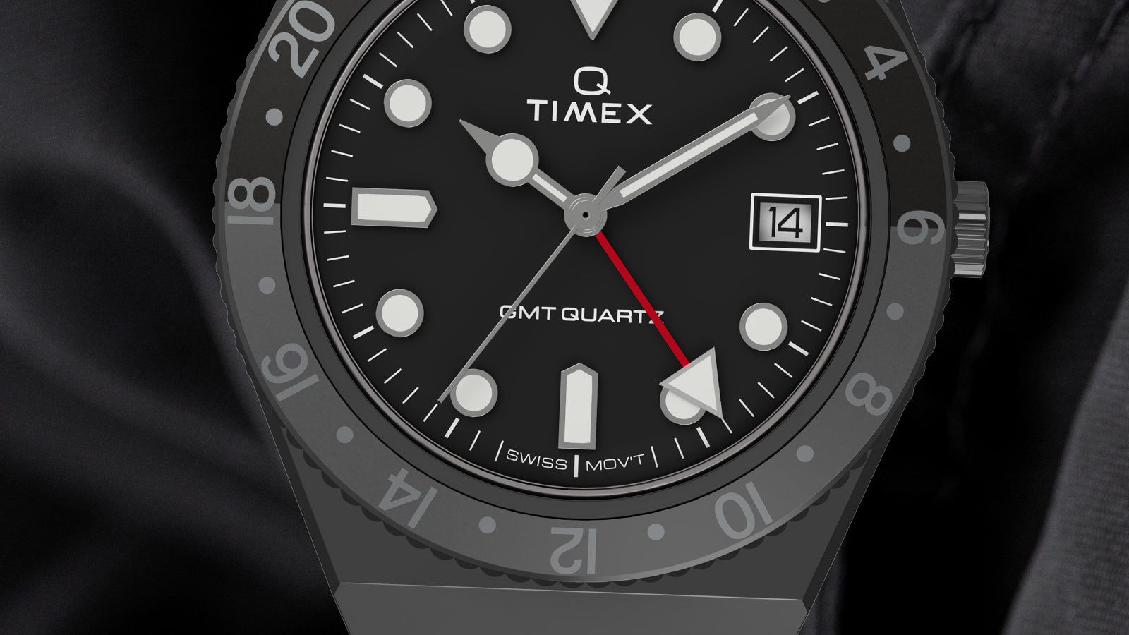 Macro shot of the Q Timex GMT dial displaying the black face with luminous white hour markers, red GMT hand, date window, and grey 24-hour bezel. The text ‘Q Timex,’ ‘GMT Quartz,’ and ‘Swiss Mov’t’ appear on the dial.