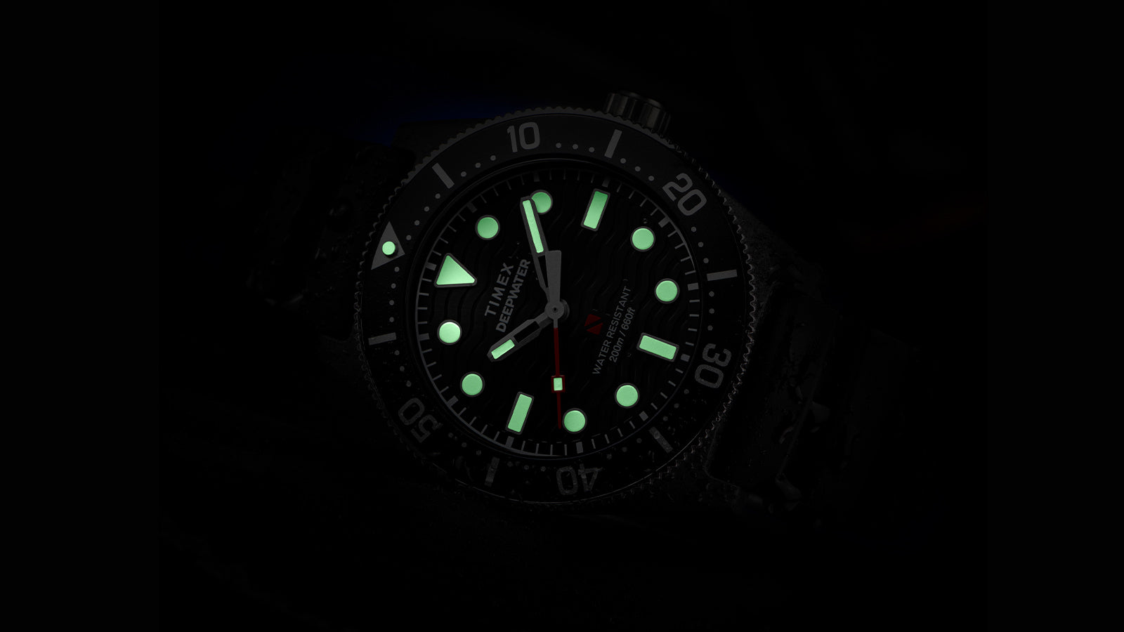 Timex Deepwater dive watch glowing in the dark, showing bright green luminescent hour markers and hands against the black dial, with the red seconds hand and bezel numerals faintly visible.