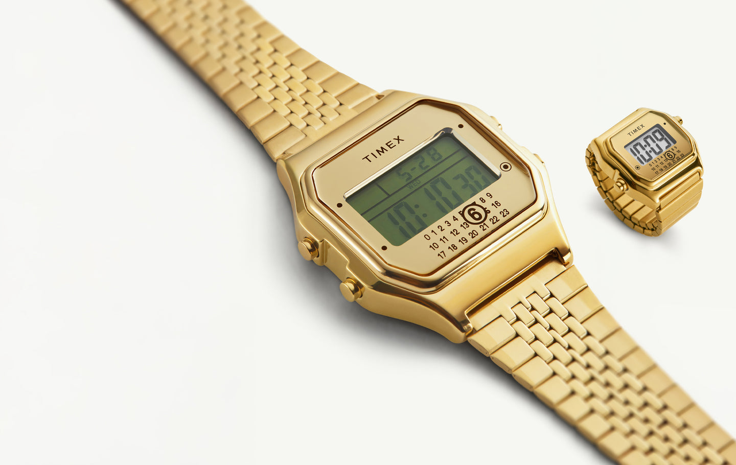 A close-up of two gold-tone digital timepieces from the Timex x MM6 Maison Margiela collection, featuring the signature Timex T80 digital watch and matching miniature ring watch. Both pieces display a retro digital screen with date and time on a sleek gold metal bracelet and ring band, set against a light background.
