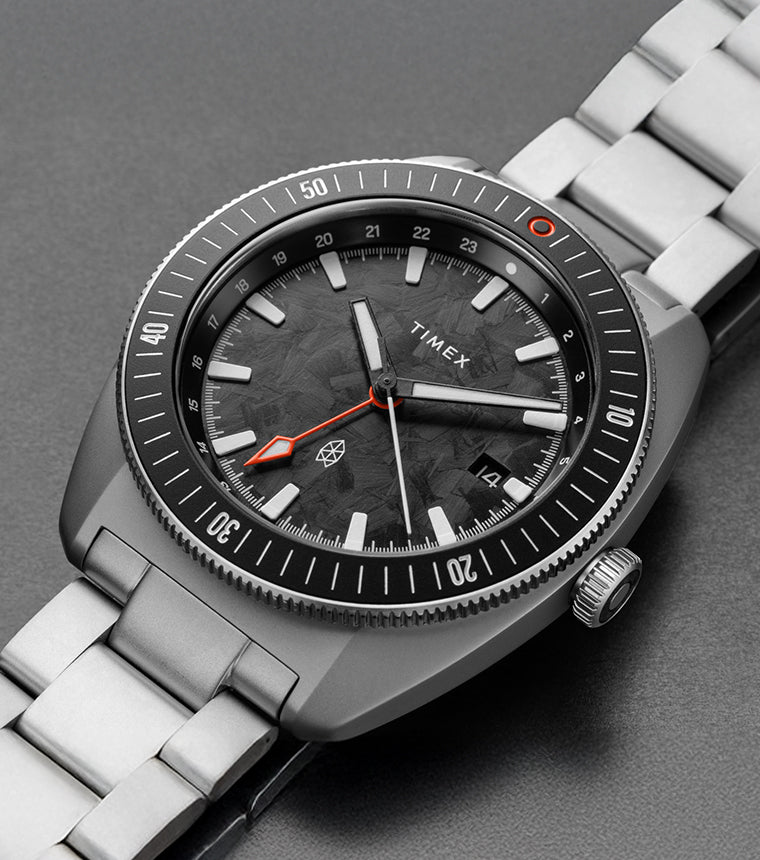 Close-up view of The James Brand x Timex GMT watch showing the black marbled dial, orange GMT hand, and stainless steel bracelet.