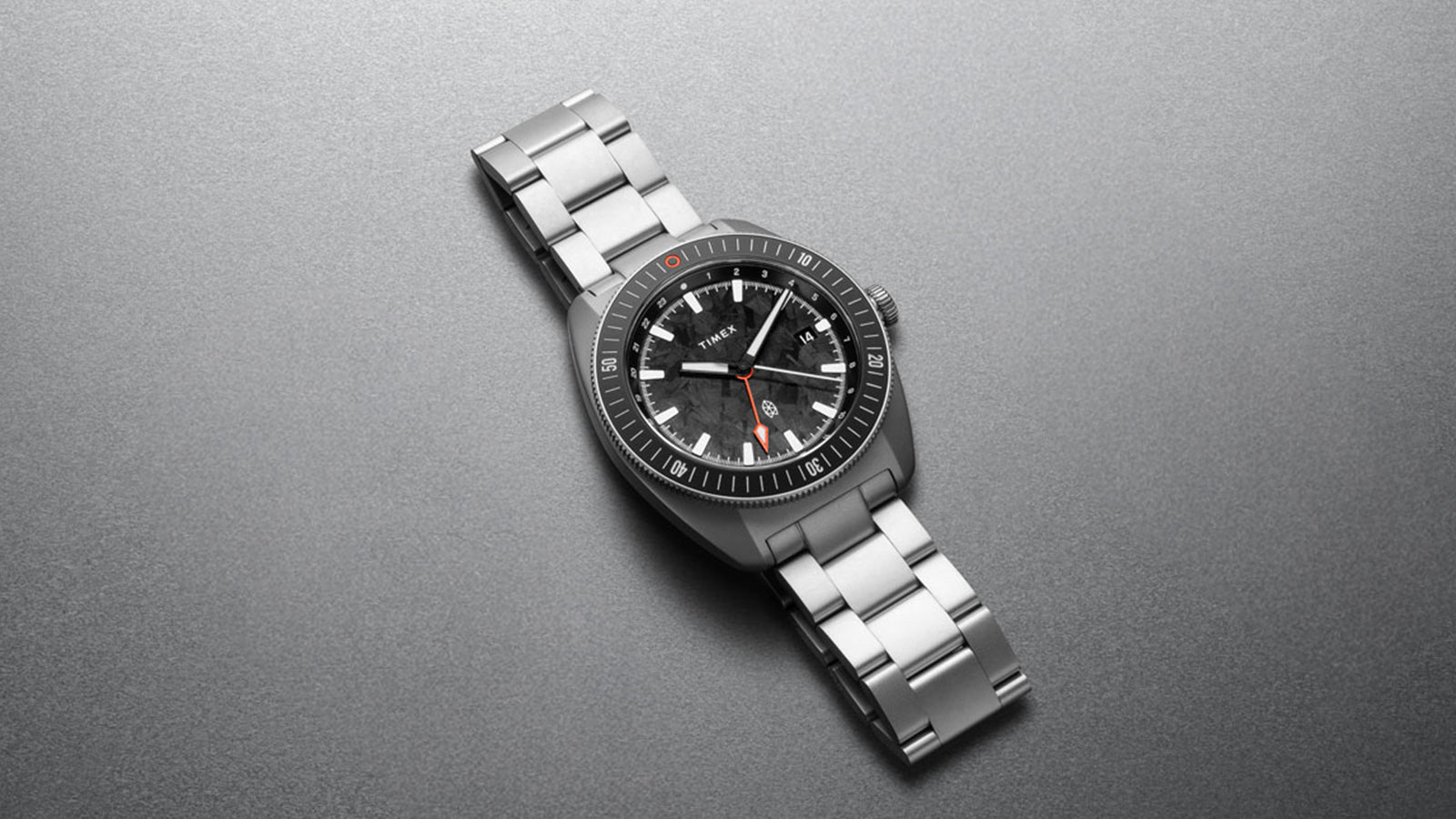 Flat lay of The James Brand x Timex GMT watch with stainless steel bracelet against a gray textured background.