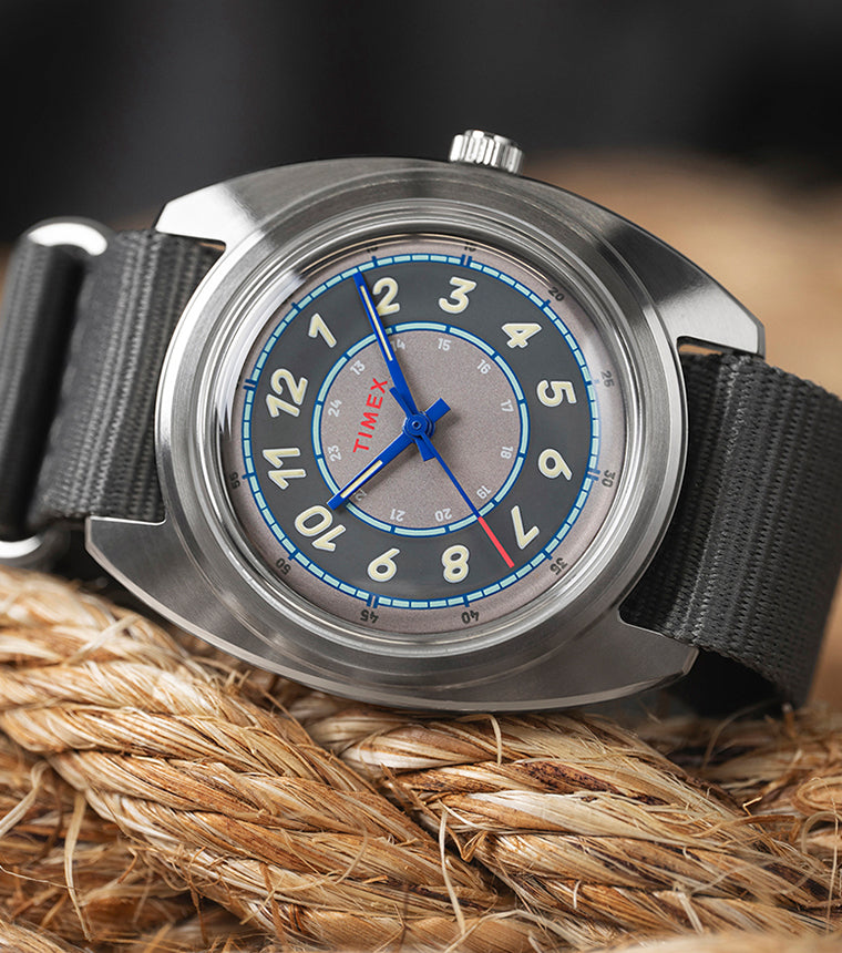 A Timex x Worn & Wound Storm watch with a gray, blue, and silver multi-layered dial resting on thick coiled rope. The watch features cream numerals, blue hands, and a gray fabric strap.
