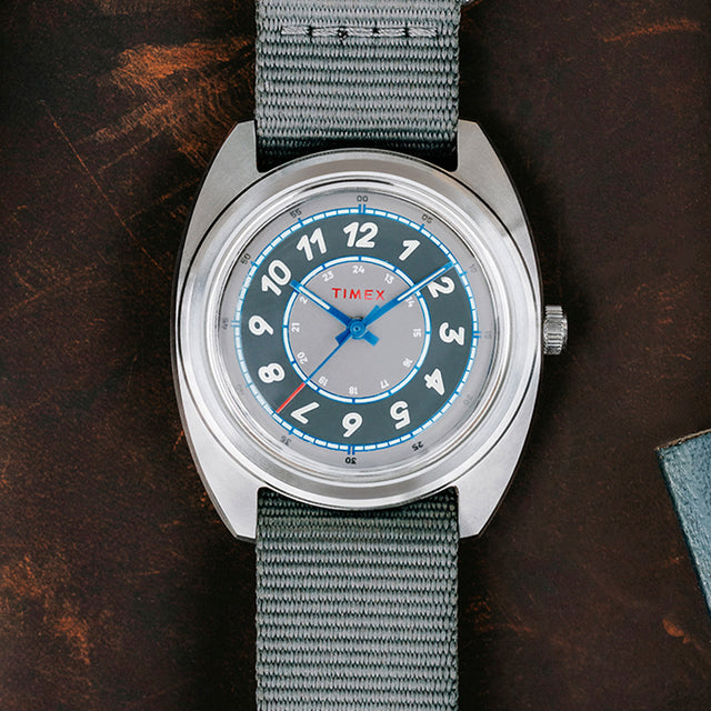 A top-down close-up of the Timex x Worn & Wound Storm watch showcasing its gray outer ring, silver center dial, bold cream numerals, and blue accents on a gray fabric strap.