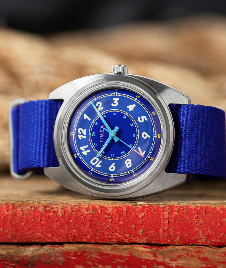 Worn & Wound x Timex Storm watch with a blue dial, bold white numerals, a brushed silver-tone case, and a blue fabric strap, resting on weathered red wood.
