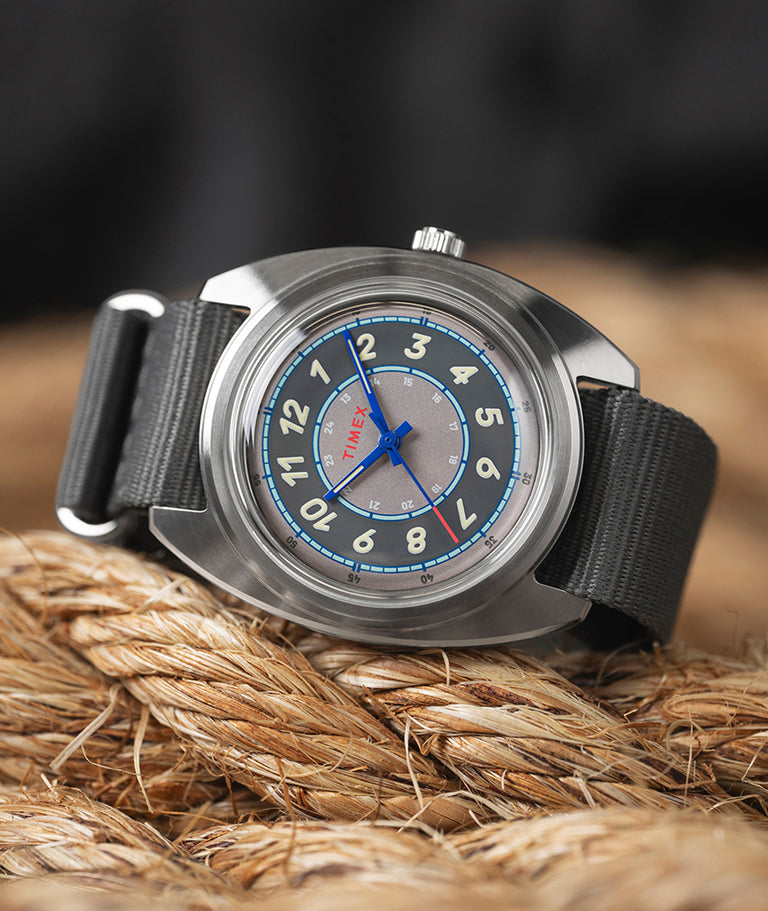 A close-up of a Timex x Worn & Wound wristwatch resting on coiled rope. The watch features a brushed stainless-steel case, a gray and blue multi-layered dial with bold cream numerals, blue hands, and a gray fabric strap. The background is softly blurred, highlighting the watch’s details.