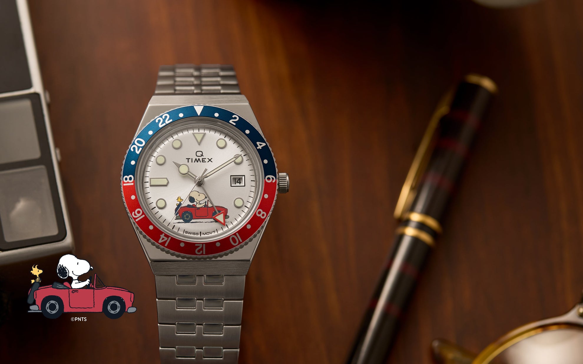 A Timex x Peanuts® Q Timex® GMT watch with a silver-tone stainless-steel bracelet and a red-and-blue 24-hour bezel displayed on a wooden desk. The silver dial features Snoopy driving a red convertible with Woodstock in the passenger seat, adding a whimsical touch to the retro GMT design.