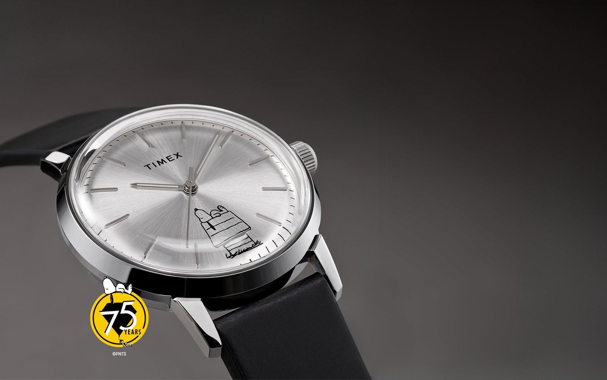A Timex x Peanuts® Marlin® Automatic 75th Anniversary Edition watch with a silver sunburst dial and black leather strap displayed on a gray background. The dial features Snoopy lounging atop his doghouse in minimalist black line art, paired with the Peanuts 75th anniversary logo.