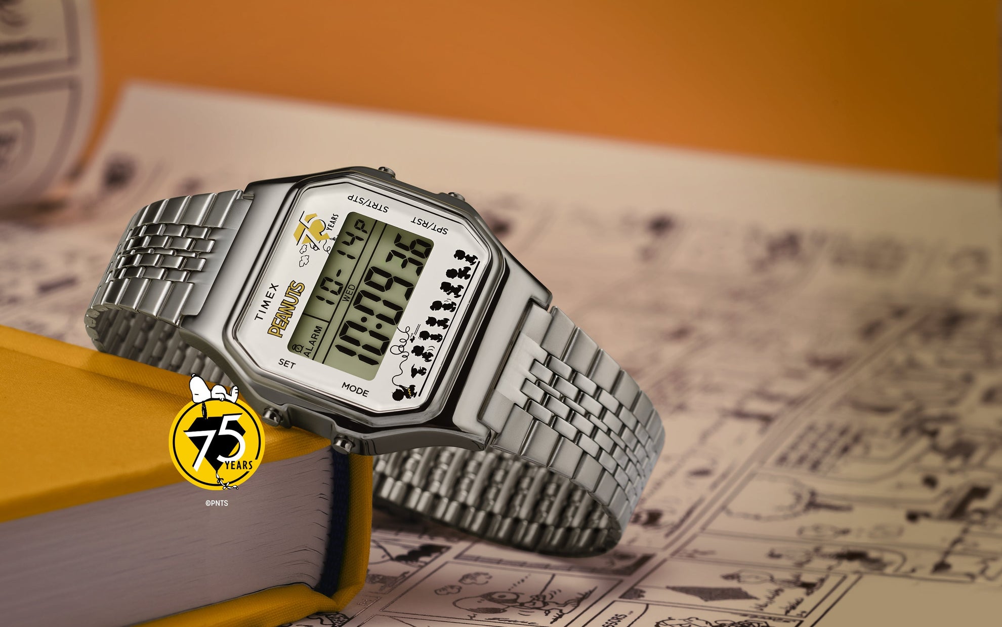 A Timex x Peanuts® T80 75th Anniversary Edition digital watch with a silver-tone stainless-steel bracelet resting on a yellow book. The display features the Peanuts 75 Years logo and silhouettes of the Peanuts characters walking along the bottom edge, set against a background of comic strip pages.