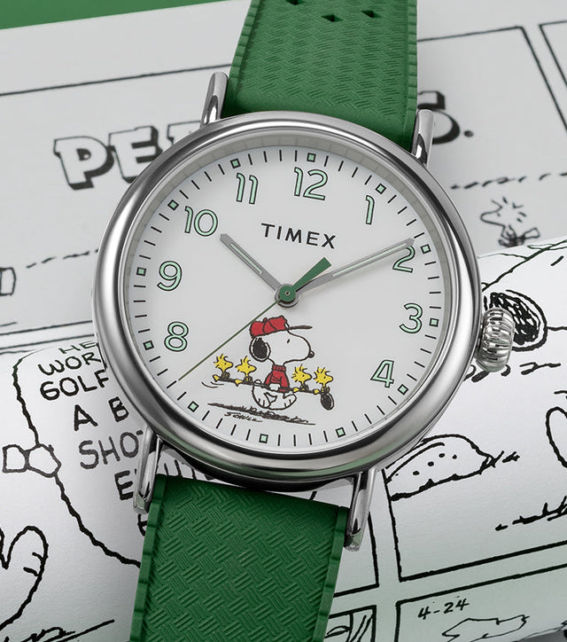 A Timex x Peanuts® Standard watch with a white dial and green strap rests on Peanuts comic strips. The dial features Snoopy dressed as a golfer in a red cap with three Woodstocks carrying golf clubs, blending sporty nostalgia with playful Peanuts charm.
