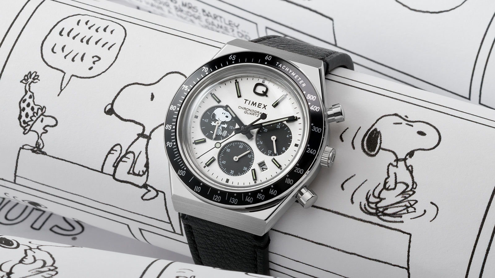 A Q Timex Chronograph x Peanuts Snoopy watch with a white dial, black sub-dials, and a black tachymeter bezel resting on Peanuts comic strips. The dial features Snoopy in one of the chronograph sub-dials, blending playful illustration with vintage racing-inspired design.
