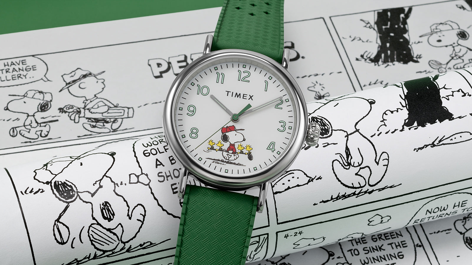A Timex x Peanuts® Standard watch with a white dial and green strap rests on Peanuts comic strips. The dial features Snoopy dressed as a golfer in a red cap with three Woodstocks carrying golf clubs, blending sporty nostalgia with playful Peanuts charm.