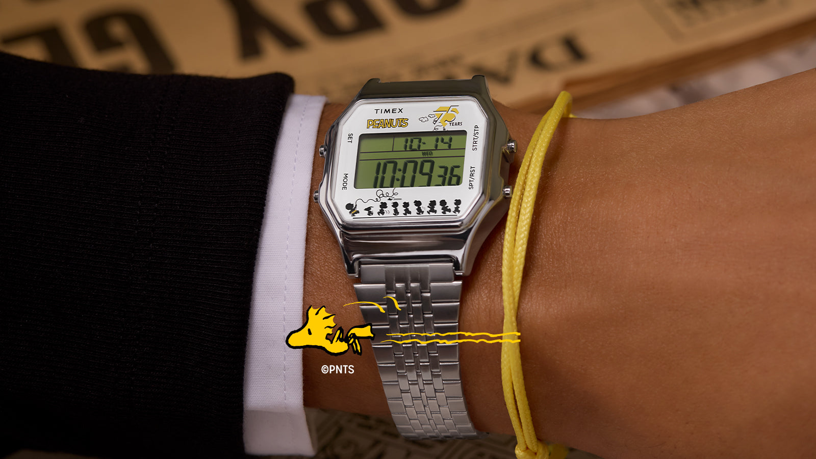 The Timex x Peanuts® T80 75th Anniversary Edition watch worn on a wrist, featuring its stainless-steel expansion band and digital display with the Peanuts 75 Years logo. The bottom of the screen shows Charlie Brown, Snoopy, and friends walking in a row, with Woodstock flying overhead.
