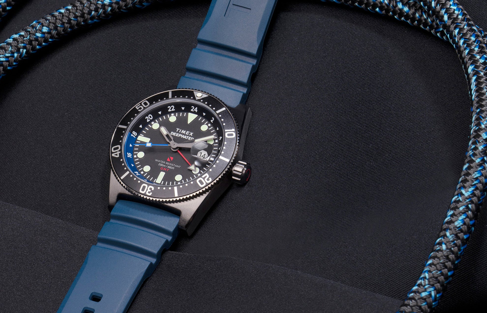 Timex Deepwater Reef 200 GMT dive watch with blue strap and black dial featuring a blue and black bezel, photographed against dark neoprene fabric and matching rope detailing.
