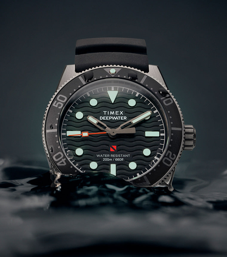 Timex Deepwater Reef 200 dive watch emerging from dark water, featuring a black dial with luminous markers and hands, black rubber strap, and 200-meter water resistance rating.