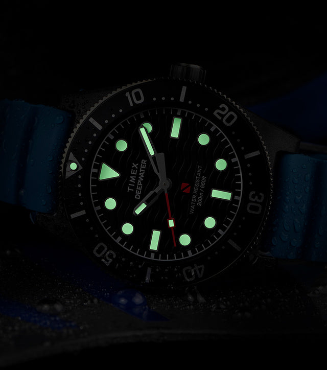 A Timex Deepwater dive watch with a black dial and luminous green markers glowing in the dark, featuring a blue silicone strap and water droplets on the case, emphasizing its water-resistant design.