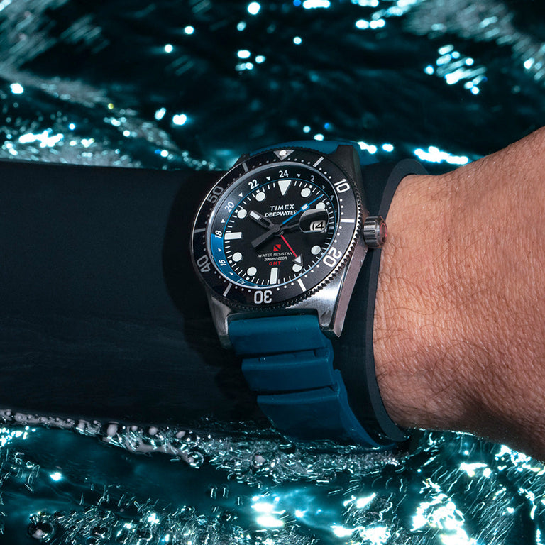 Diver wearing the Timex Deepwater Reef 200 watch underwater, the blue strap and stainless steel case reflecting light as bubbles rise around them.