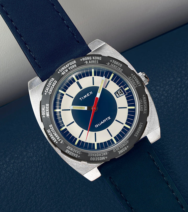 Top-down view of the Q Timex® 1972 World Time watch with navy blue strap and blue-and-cream dial resting on blue textured leather.