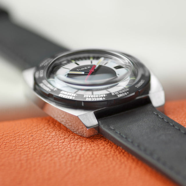 Low side view of the Timex World Time Reissue watch highlighting the curved crystal dome, brushed steel case edges, and black leather strap against an orange leather background.