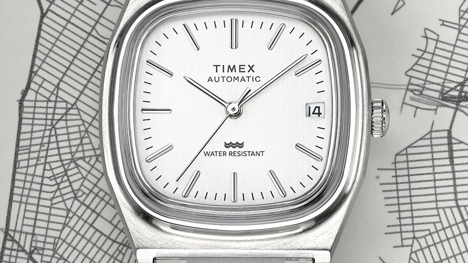 Detailed view of the silver dial of the Timex® Automatic 1983 E Line Reissue featuring minimalist baton indices, slim hands, a date window at 3 o’clock, and the TIMEX Automatic logo.