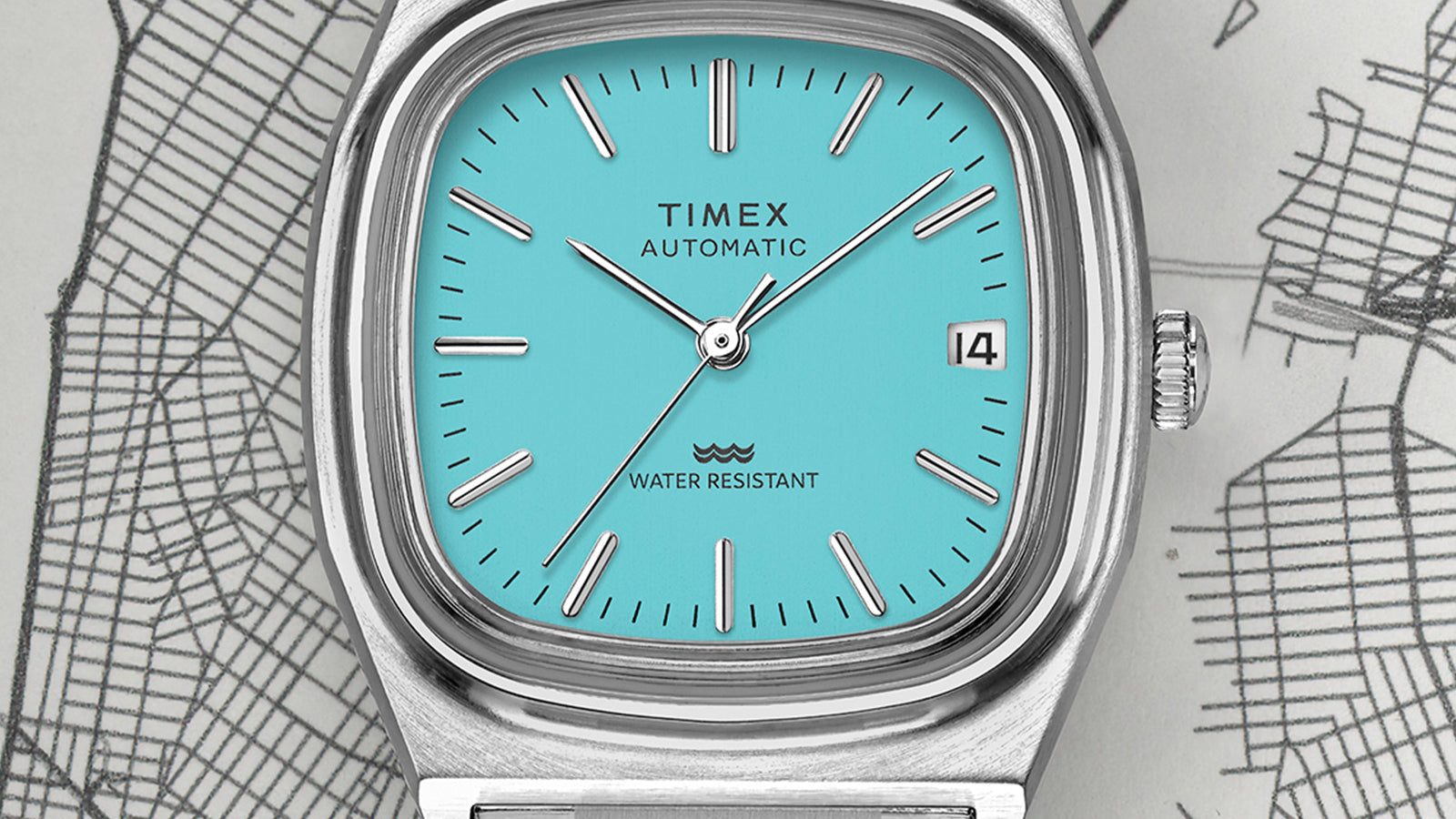 Close-up of the turquoise dial of the Timex® Automatic 1983 E Line Reissue, highlighting minimalist baton hour markers, a date window at 3 o’clock, and silver-tone hands.