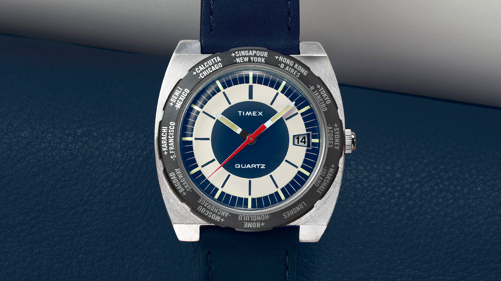 Top-down view of the Q Timex® 1972 World Time watch with navy blue strap and blue-and-cream dial resting on blue textured leather.