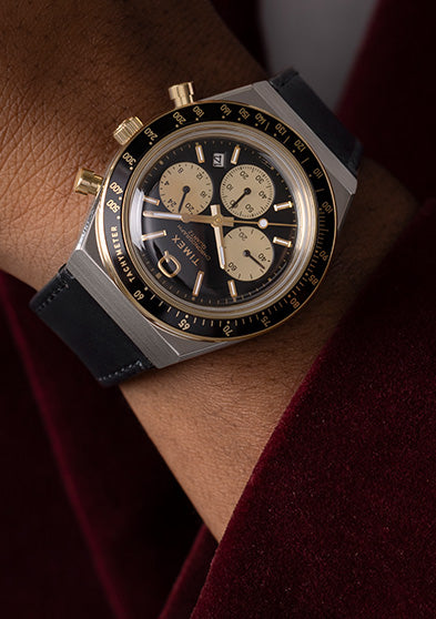 Q Timex Chronograph with black and gold dial worn on wrist, paired with a dark red jacket, showcasing a refined vintage-inspired look.
