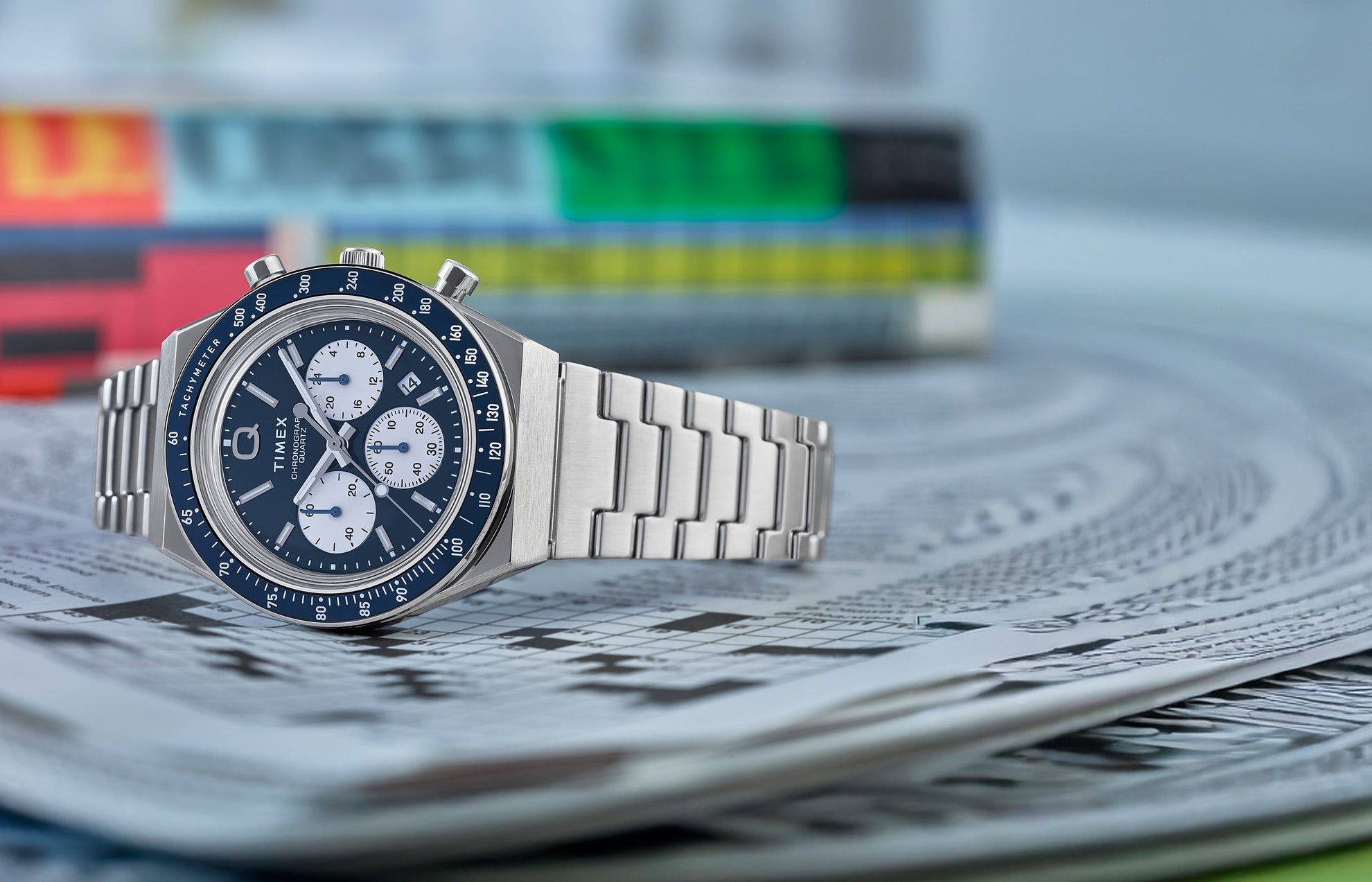 Q Timex Chronograph with a blue dial, white sub-dials, and stainless steel bracelet resting on a folded newspaper with colorful pages in the background.