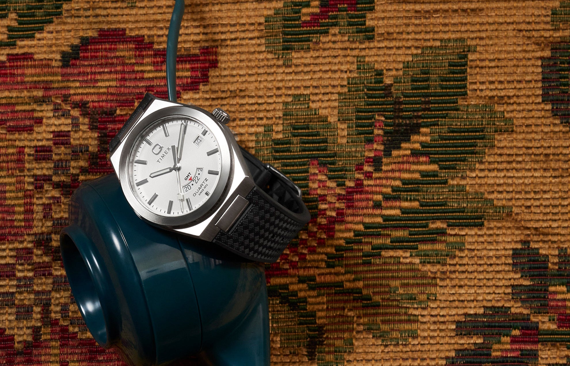 Q Timex® Continental GMT watch with a silver dial, stainless-steel case, and black woven strap displayed on a vintage tapestry background beside a teal rotary phone.