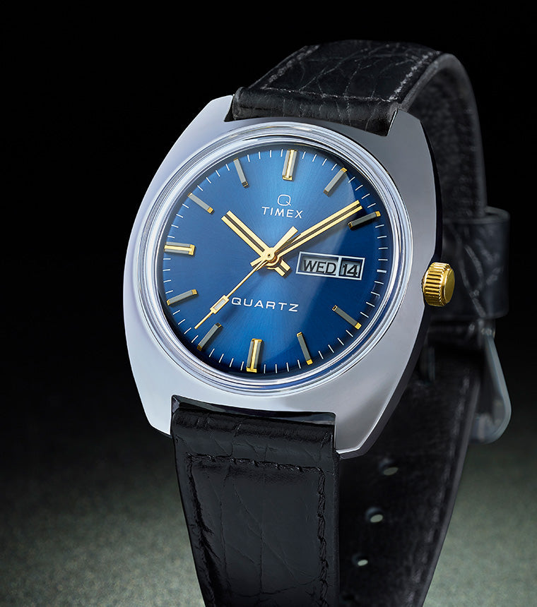 Q Timex® 1978 Day/Date watch with a blue sunray dial, gold-tone hands, and a black leather strap, displayed against a dark background with a subtle spotlight effect.