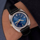 Q Timex® 1978 Day/Date watch worn on the wrist, showing the blue dial, gold-tone details, and black leather strap against a dark sleeve.