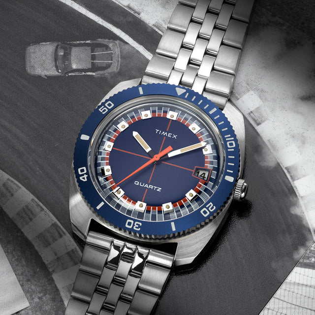 Q Timex® 1971 Velocity watch with a blue dial and silver bracelet, placed over a black-and-white racing track photo, emphasizing its vintage motorsport inspiration.
