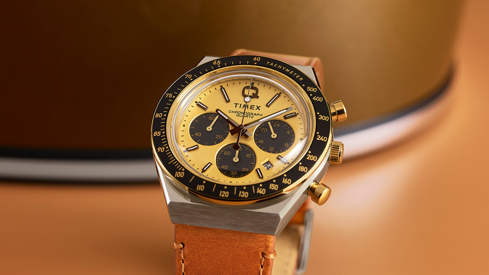 Close-up of the Q Timex Chronograph highlighting its gold dial, black sub-dials, tachymeter bezel, and gold-tone pushers and crown.