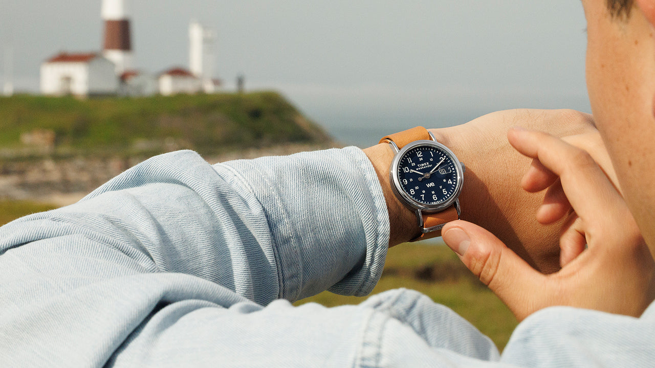 Lifestyle shot of a Timex Weekender with blue dial and brown leather strap, worn on wrist while adjusting time outdoors with a lighthouse in the background.