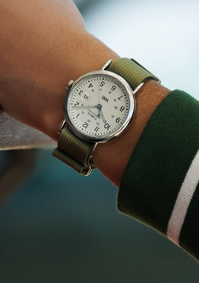 Timex Weekender watch with green strap worn on a wrist, styled casually with a green and white striped sweatshirt near sunglasses on a table.