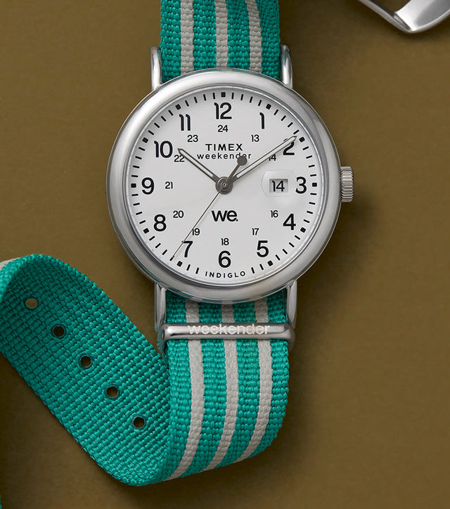 A Timex Weekender watch with a white dial, black numerals, and a teal-and-white striped fabric strap, displayed against a brown background.