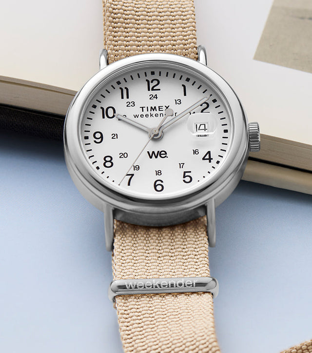 Timex Weekender watch with a beige fabric strap and white dial, placed beside an open book showing a vintage beach photograph.