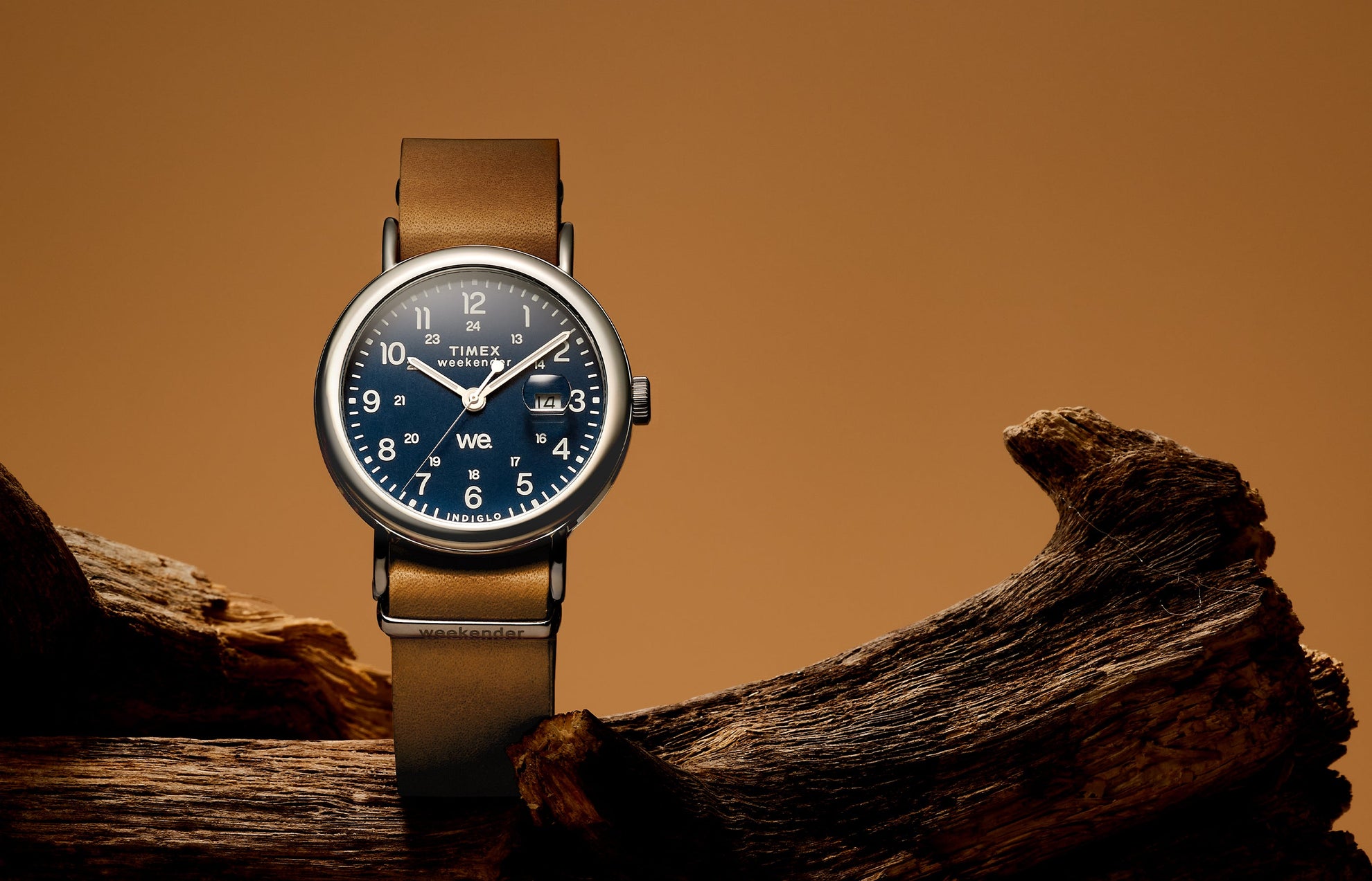 Timex Weekender watch with a blue dial, silver-tone case, and brown leather strap, displayed on driftwood with a warm brown background.